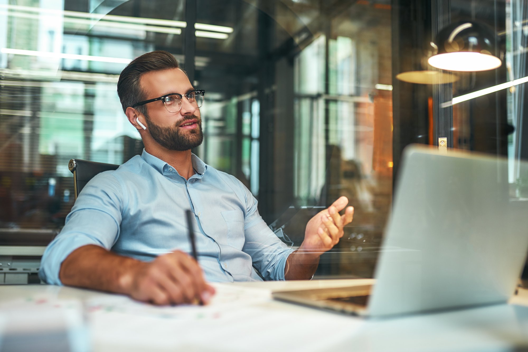 Business talk. Young and successful bearded man in eyeglasses and headphones talking with client by phone while sitting in the modern office Business talk. Young and successful bearded man in eyeglasses and headphones talking with client by phone while sitting in the modern office